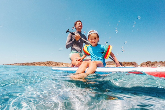 Kind mit bunten Schwimmflügeln auf Paddelboard mit Vater auf dem Meer